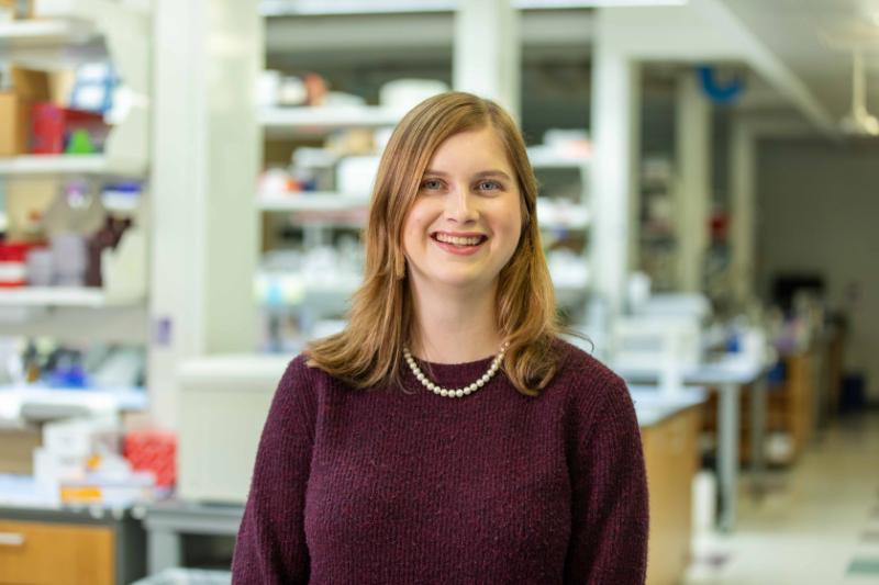 A student smiling at the camera with a lab behind her.